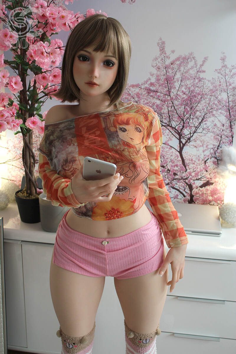 Besonders sympathische Realdoll Yelan mit rosa Shorts und langärmligem Shirt, stehende Ganzkörperansicht im Schlafzimmer
